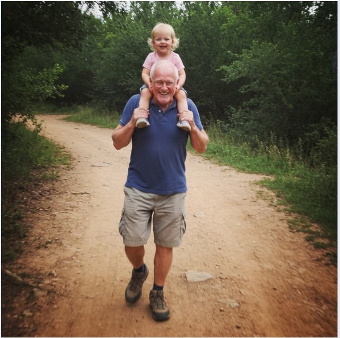 Ad creative: a gray-haired man carries a smiling toddler on his shoulders down a wooded dirt trail