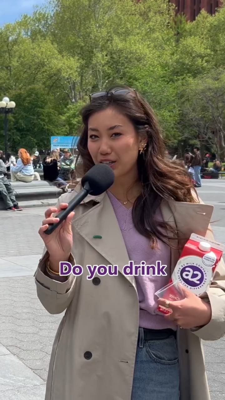In a city park, a host in a trench coat holds a mic and an a2 Milk carton, mid-question with the caption “Do you drink”