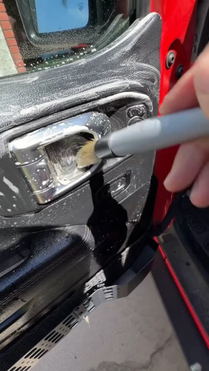 Handheld POV close-up of a detailing brush scrubbing foamy cleaner into the chrome door handle of a red Jeep's interior door panel in the sun; no on-screen text