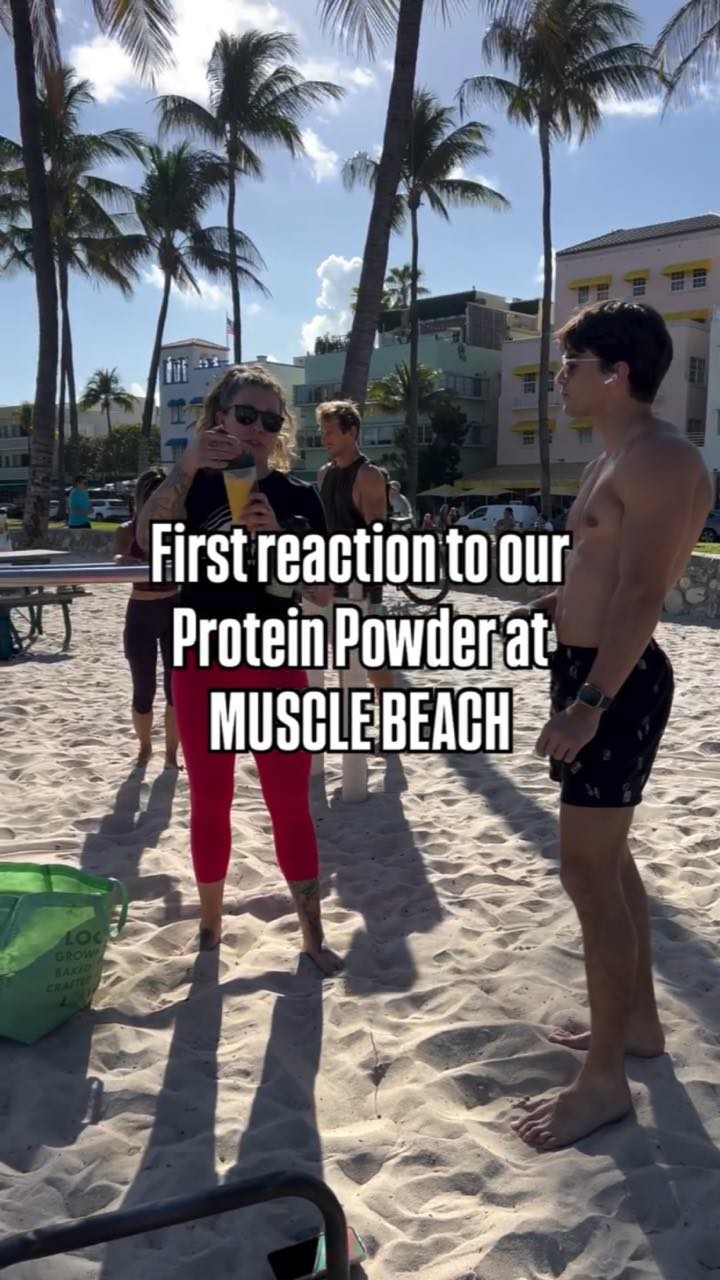 On a palm-lined beach, a woman in sunglasses sips from a sample cup beside a shirtless guy, captioned “First reaction to our Protein Powder at MUSCLE BEACH”