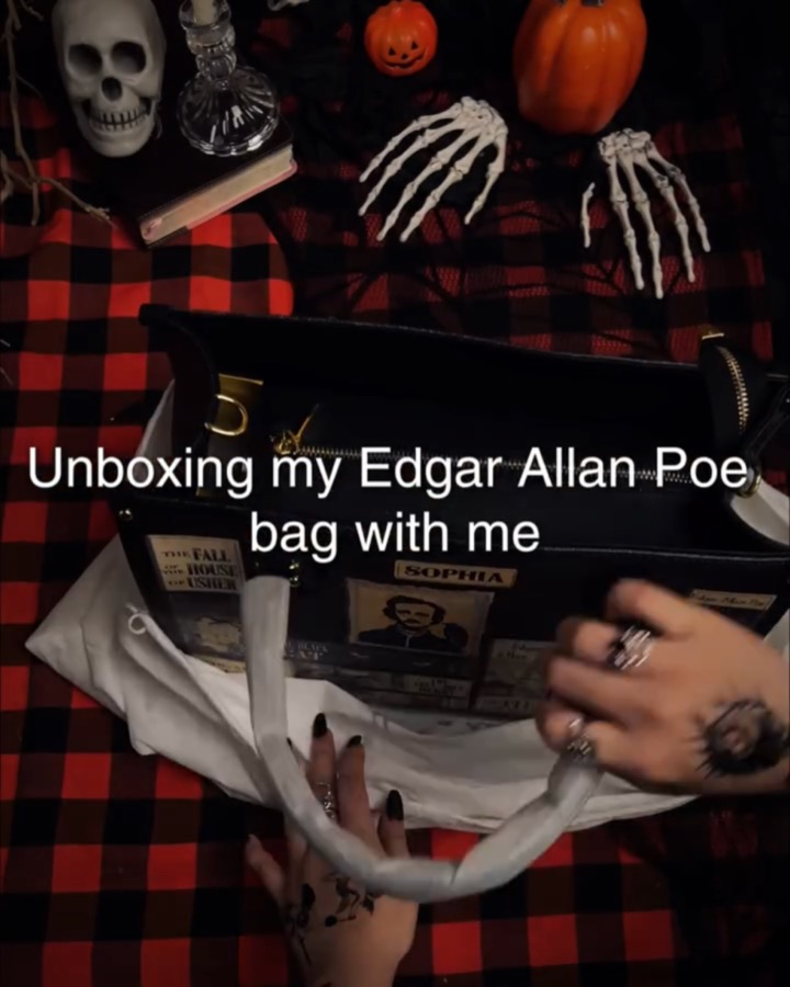 Top-down hands-only unboxing on red plaid with a skull, pumpkins and skeleton hands: tattooed hands pull a black Poe-themed handbag from its dust bag, captioned “Unboxing my Edgar Allan Poe bag with me”