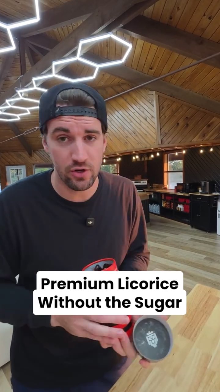 A man in a backwards cap holds an open red-and-black tub of black licorice in a wood-paneled room, captioned “Premium Licorice Without the Sugar”