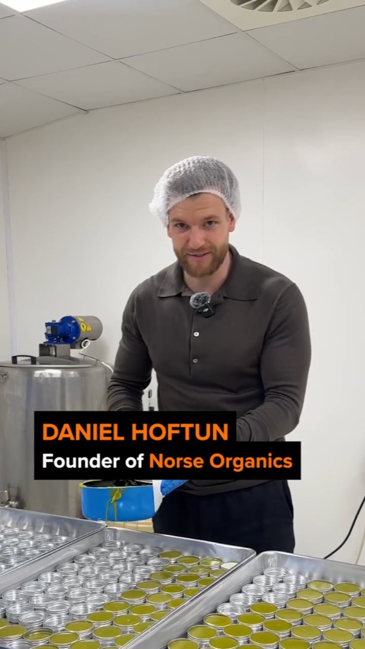 Founder in a hairnet and lav mic stands over trays of filled balm tins on a production floor, with a lower third reading “DANIEL HOFTUN Founder of Norse Organics”