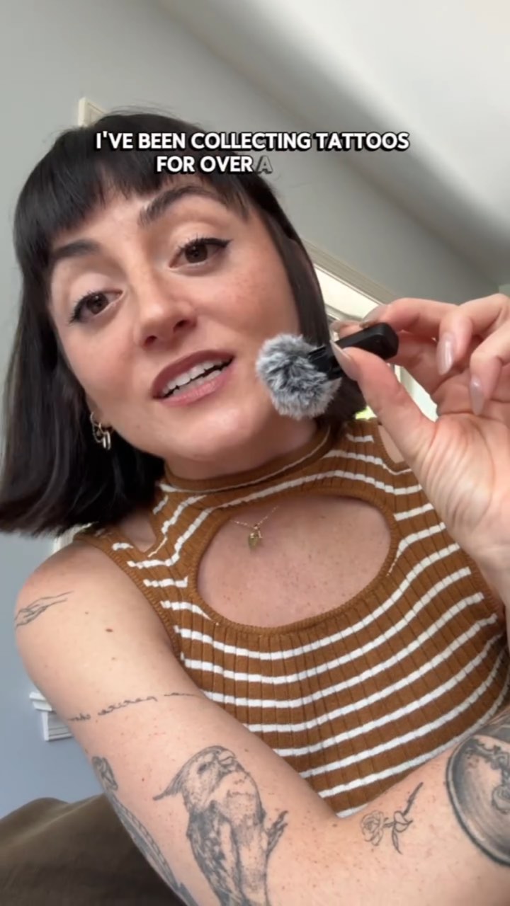 Low-angle selfie of a tattooed creator in a striped knit top holding a fuzzy lav mic, captioned “I'VE BEEN COLLECTING TATTOOS FOR OVER A…”