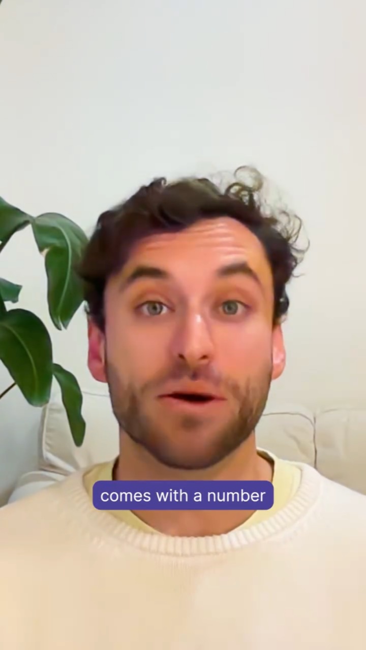 A curly-haired man in a cream sweater talks wide-eyed to camera in front of a plant, with a purple subtitle reading “comes with a number”