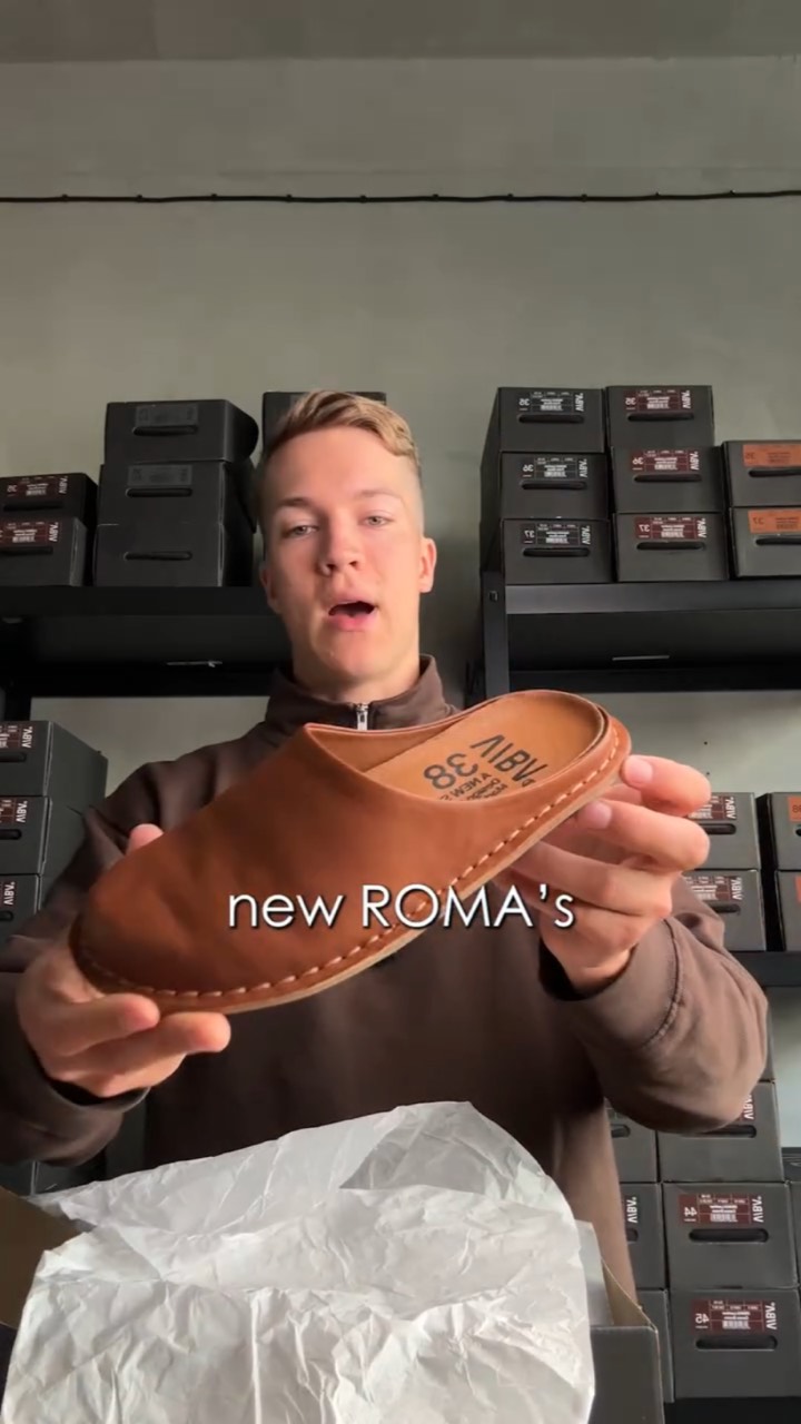 A young man in a shoe stockroom lifts a tan leather slipper out of tissue paper toward the camera, captioned “new ROMA’s”
